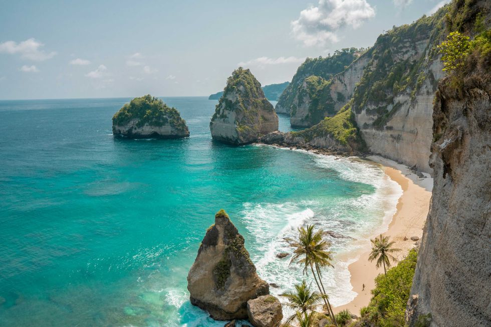 Turquoise sea with rocky cliffs and white sand beach, dotted with palm trees.