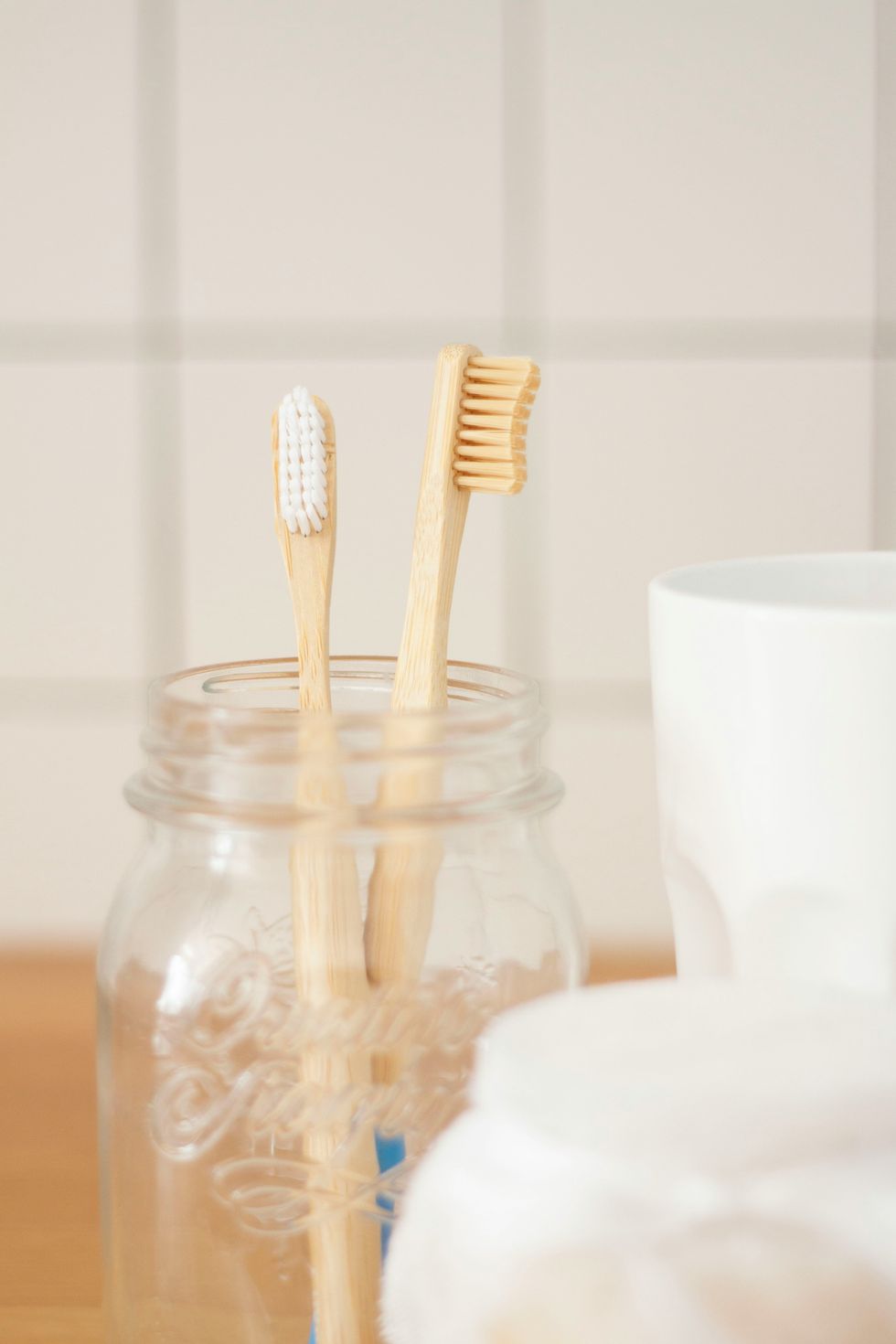 Two bamboo toothbrushes in a glass jar beside a white cup on a bathroom countertop.