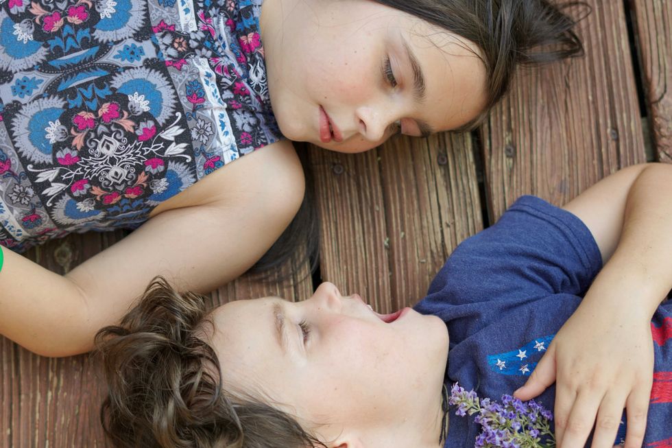 Two children lying on wooden planks, facing each other, holding small purple flowers.