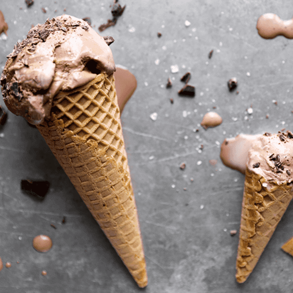 Two chocolate ice cream cones on a gray surface with scattered chocolate pieces.