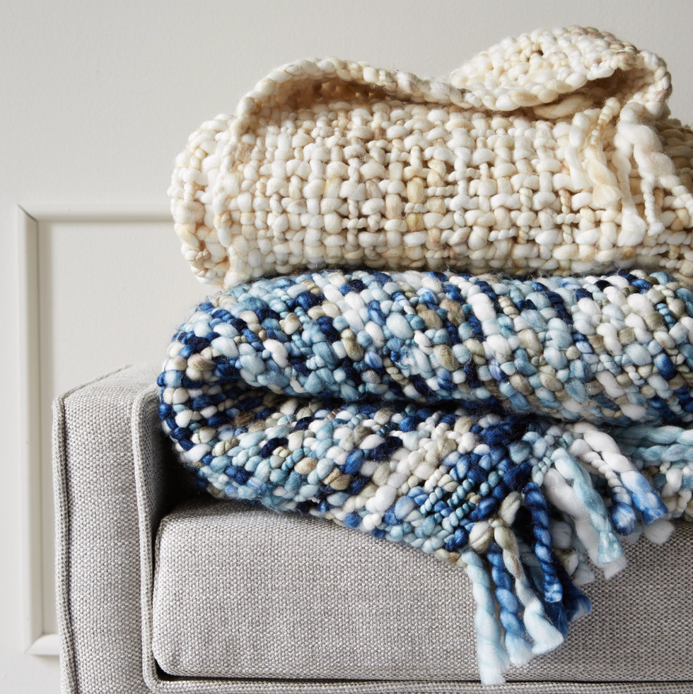 Two chunky knit blankets, one beige and one blue, folded on a gray sofa arm.