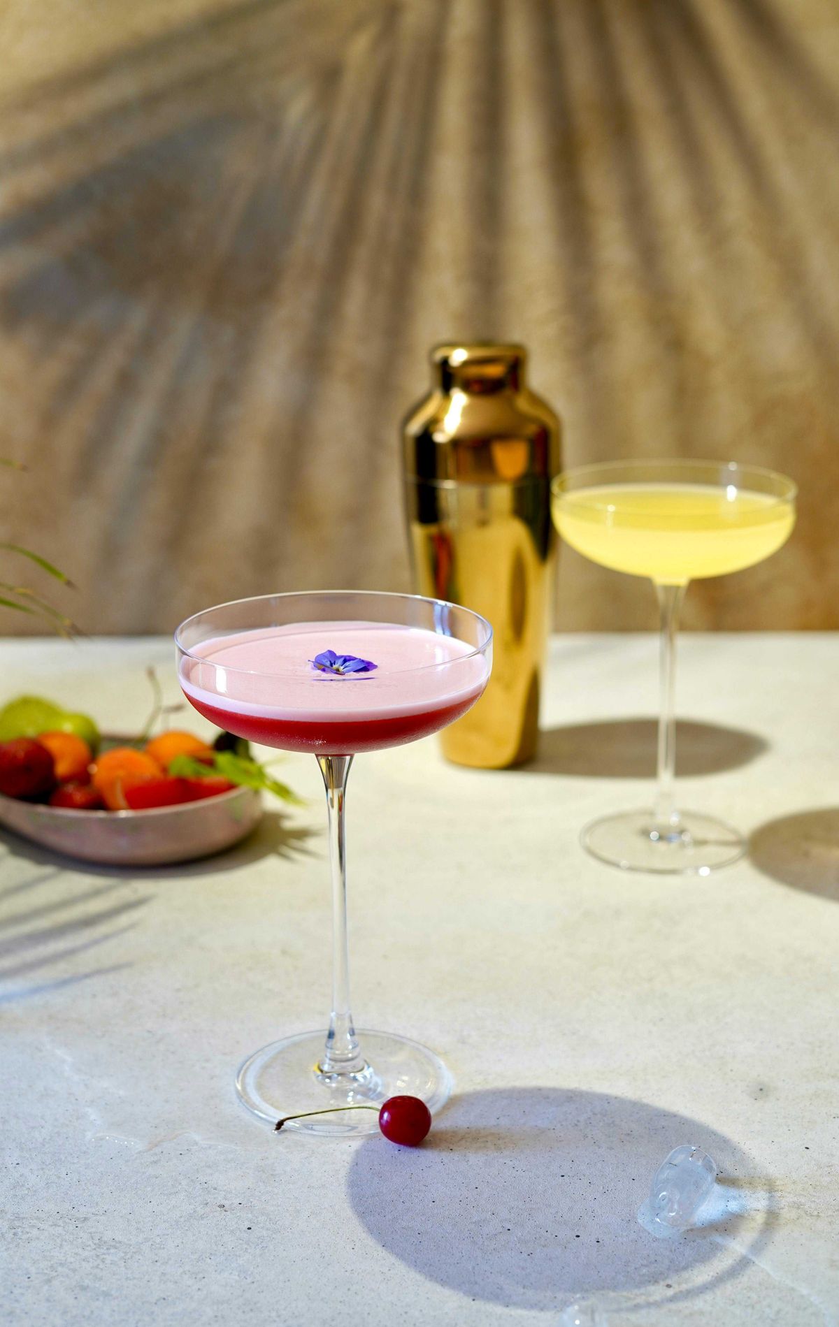 Two cocktails on a table with a gold shaker and fruit in the background.