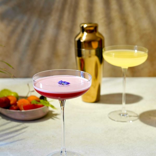 Two cocktails on a table with a gold shaker and fruit in the background.