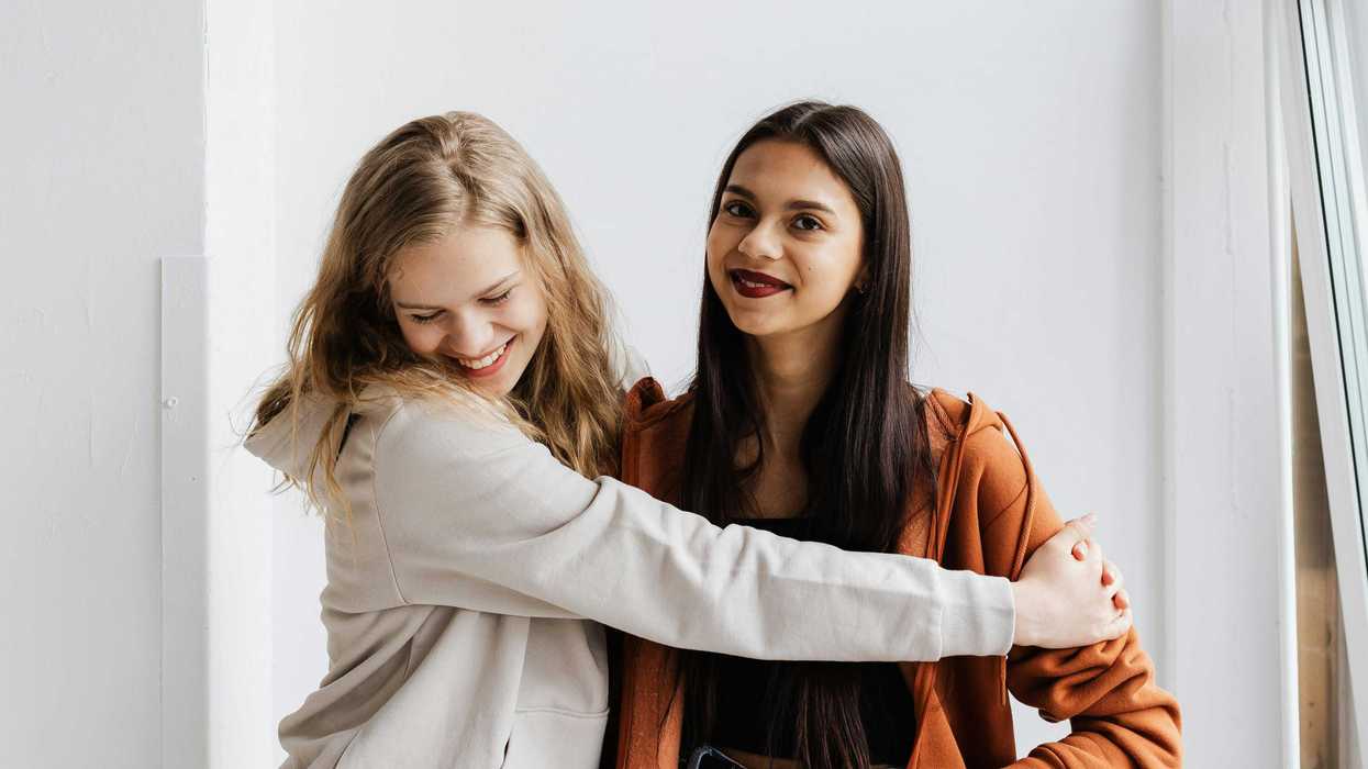 Two friends smiling, one in white and one in orange hoodies, posing in a bright room.