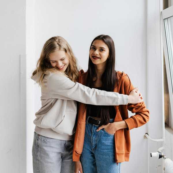 Two friends smiling, one in white and one in orange hoodies, posing in a bright room.