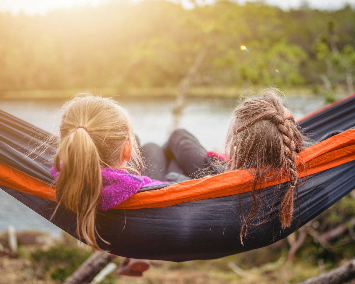 Two girls relax in a hammock by a serene lake, surrounded by nature.