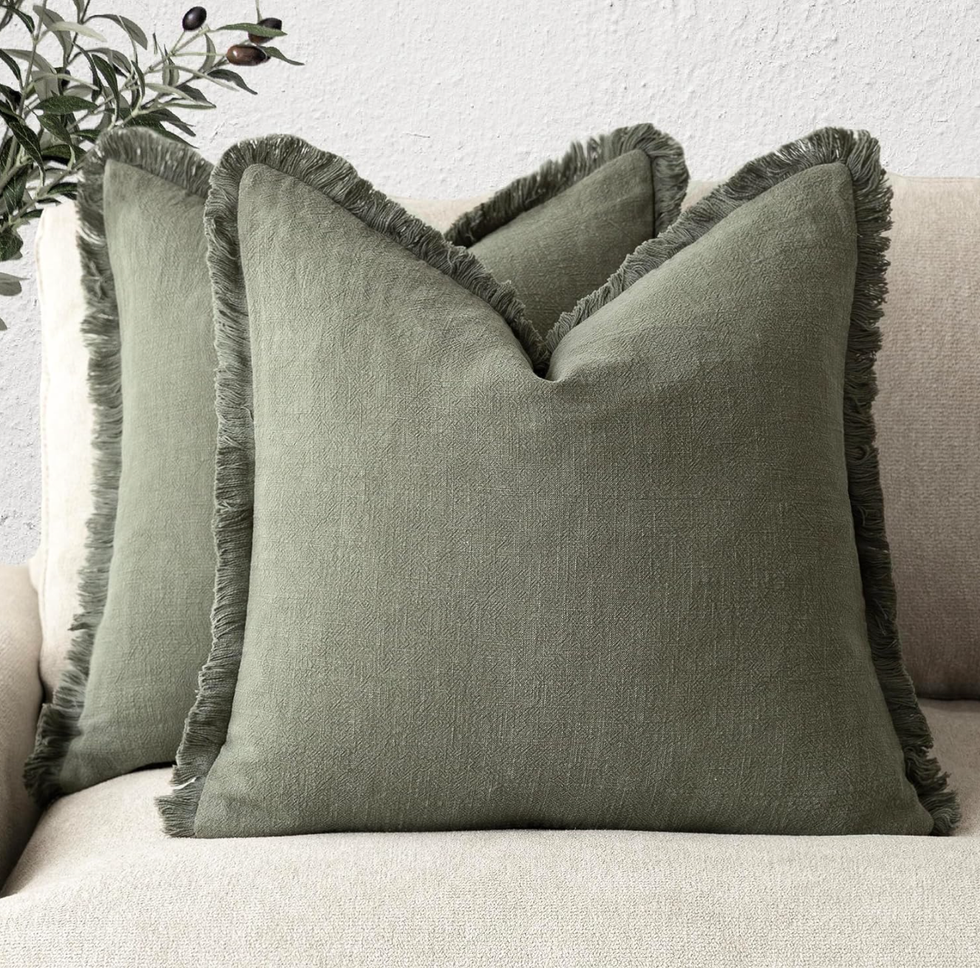 Two green fringed pillows on a beige sofa with leafy decor in the background.