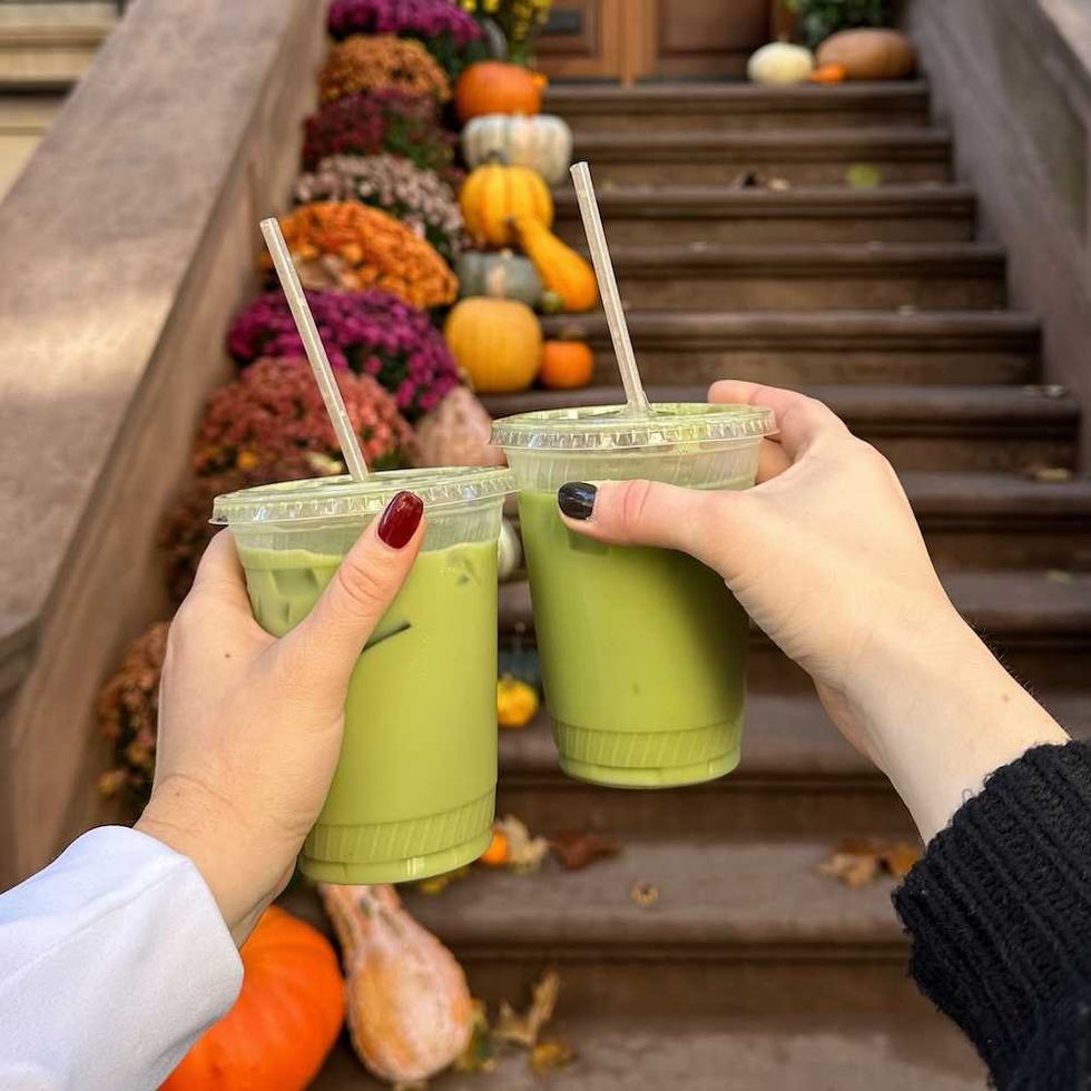 Two hands holding green drinks in front of steps with pumpkins and flowers.