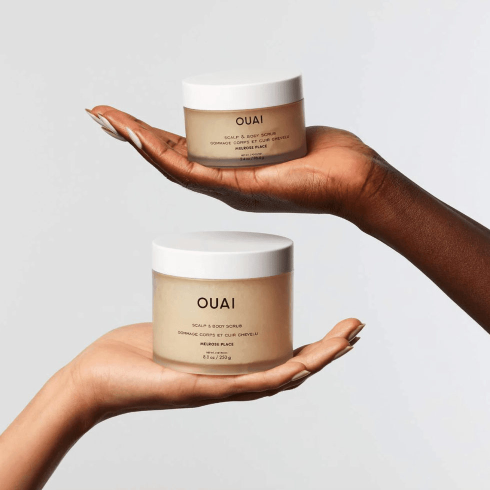 Two hands holding jars of Ouai Scalp & Body Scrub against a plain background.