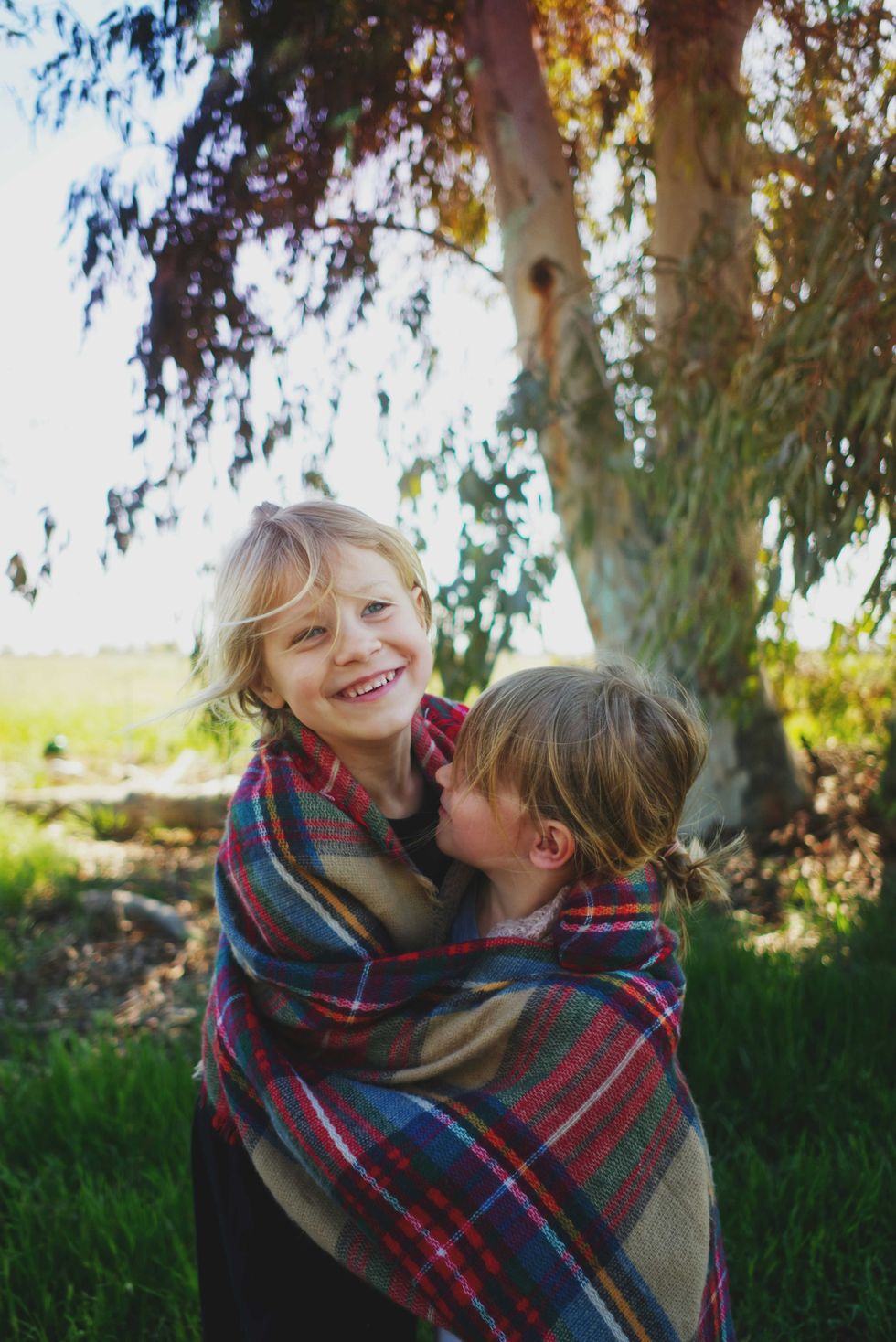 Two kids wrapped in a plaid blanket, smiling, standing under a tree.