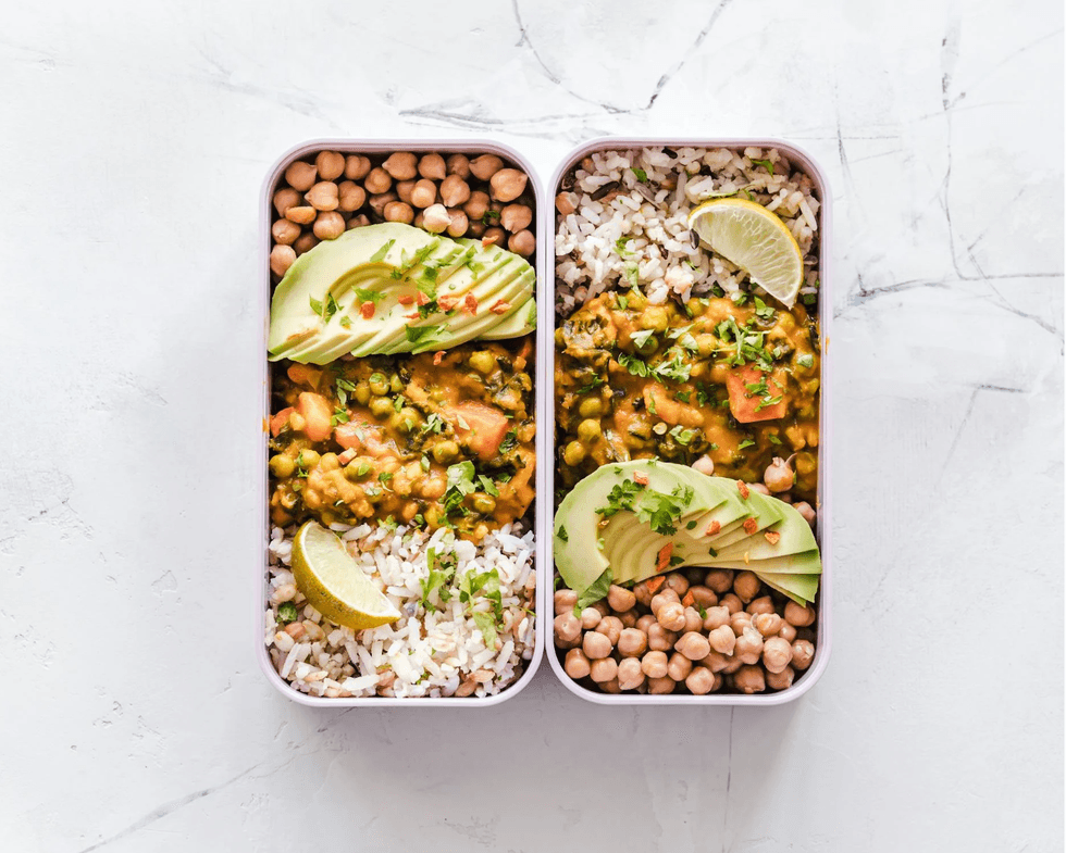 Two lunchboxes with rice, curry, chickpeas, avocado slices, and lime wedges on a light background.