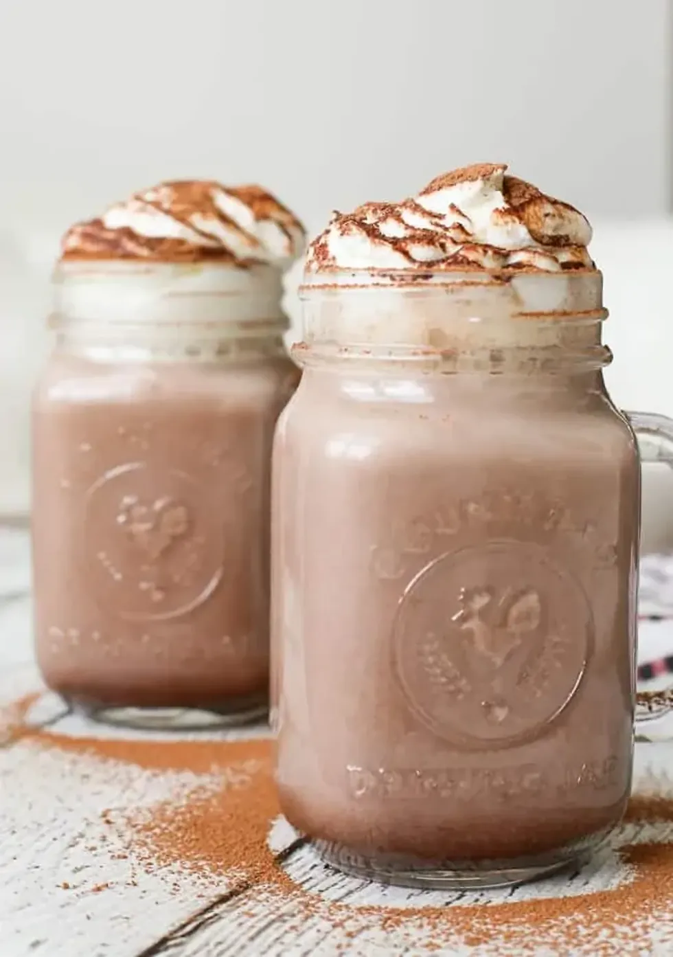 Two mason jars of hot cocoa topped with whipped cream and cocoa powder.