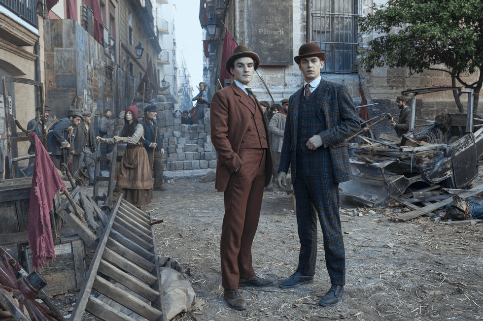 Two men in vintage suits stand in a historical scene with a bustling street and makeshift barricades.