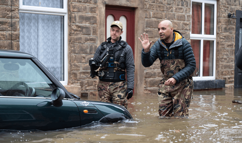 Two men in waders navigate knee-deep floodwaters beside a partially submerged car.