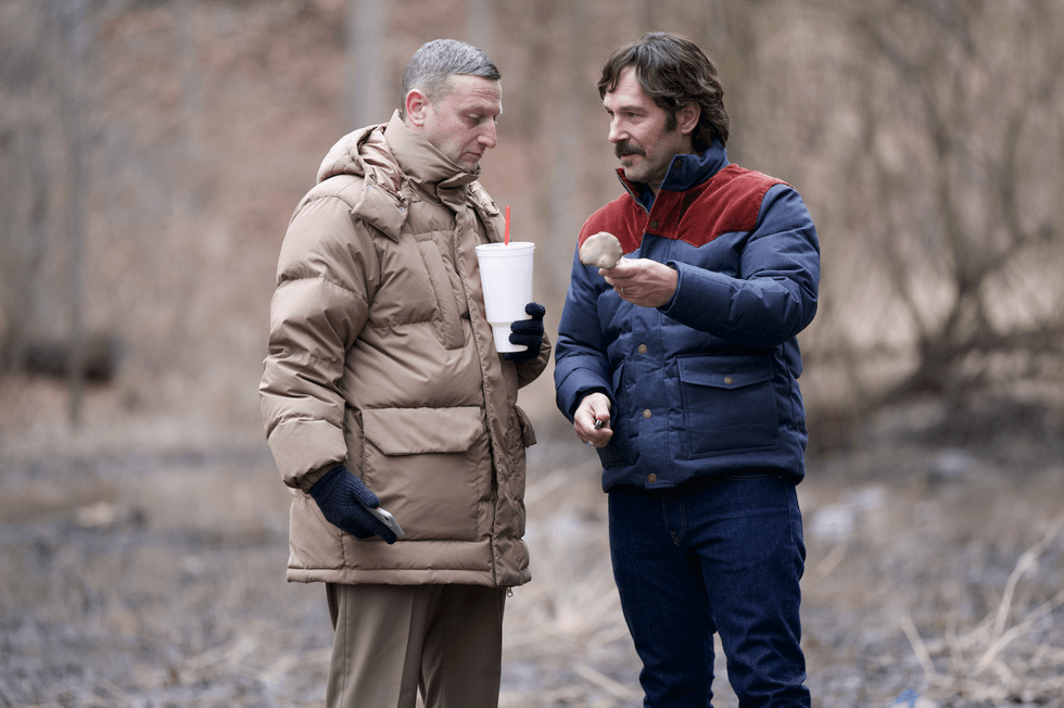 Two men in winter coats stand outside, one holding a drink, the other showing a mushroom.