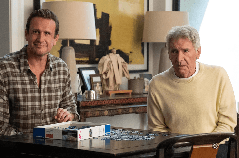 Two men sit at a table working on a puzzle in a cozy room with warm lighting.