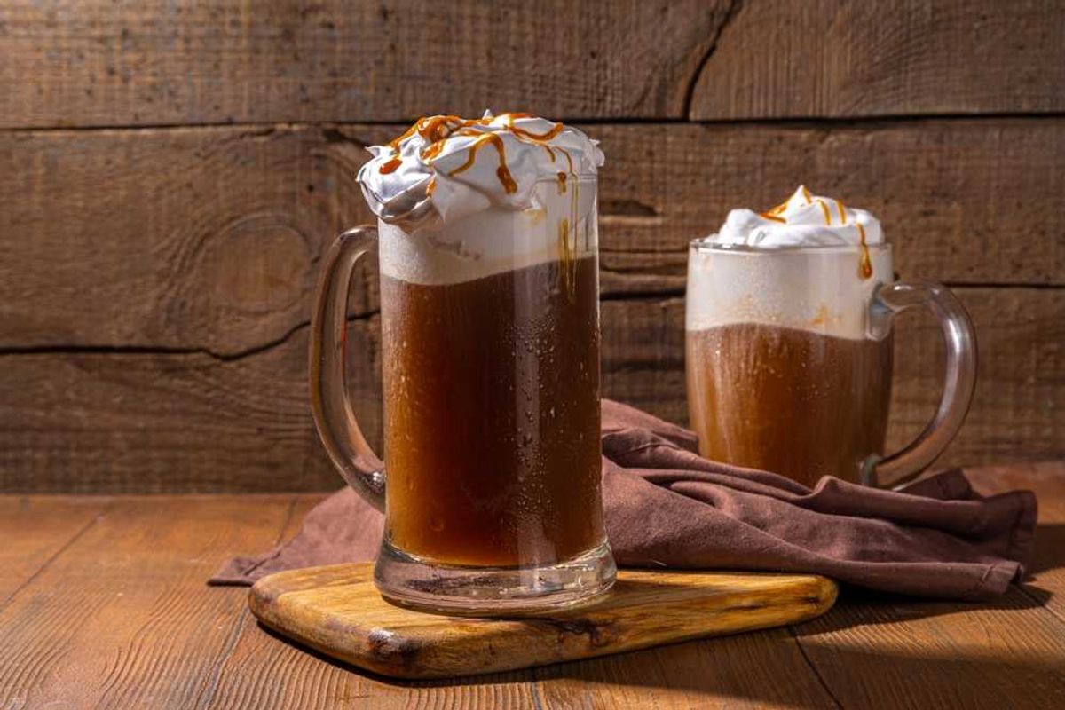 Two mugs of root beer float with whipped cream and caramel, on a wooden board.