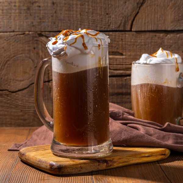 Two mugs of root beer float with whipped cream and caramel, on a wooden board.