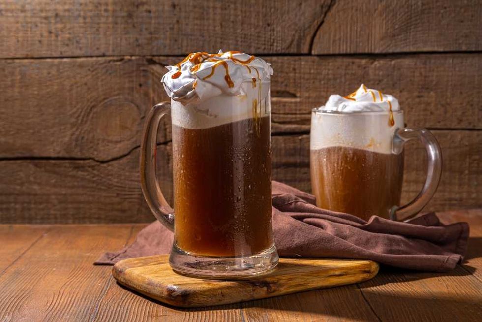 Two mugs of root beer float with whipped cream and caramel, on a wooden board.