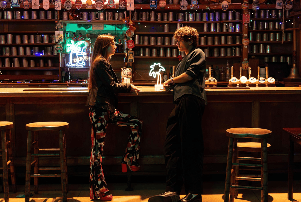 Two people chat at a dimly lit bar with neon lights and colorful d\u00e9cor.