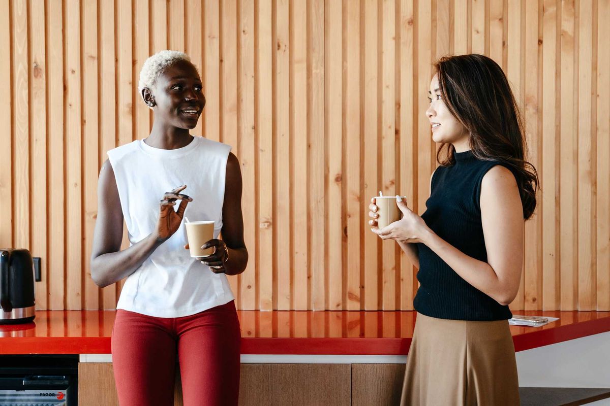 Two people chatting and holding cups in front of a wooden wall.