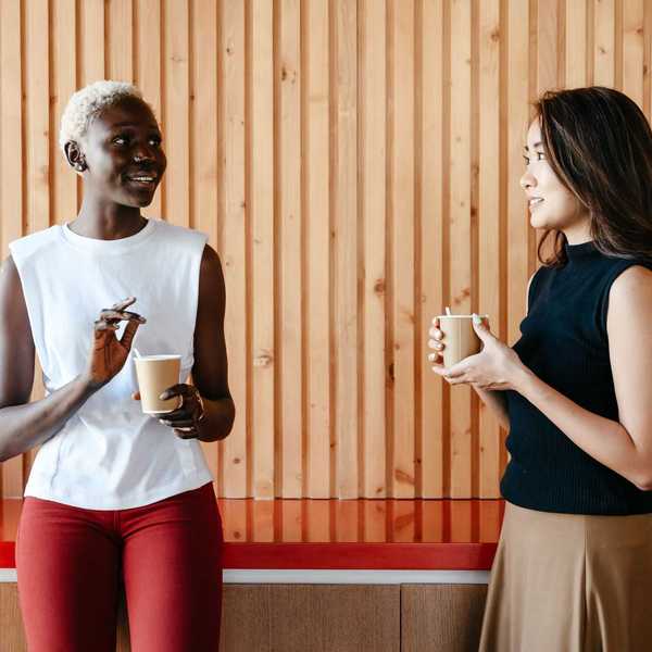 Two people chatting and holding cups in front of a wooden wall.