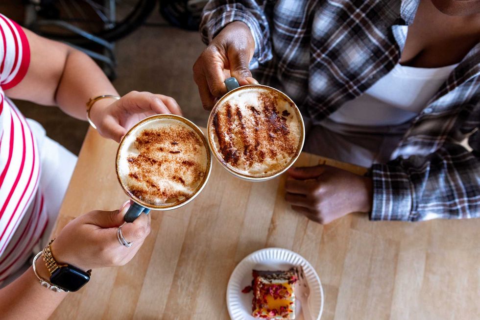 Two people clinking cinnamon lattes over a table with cake.
