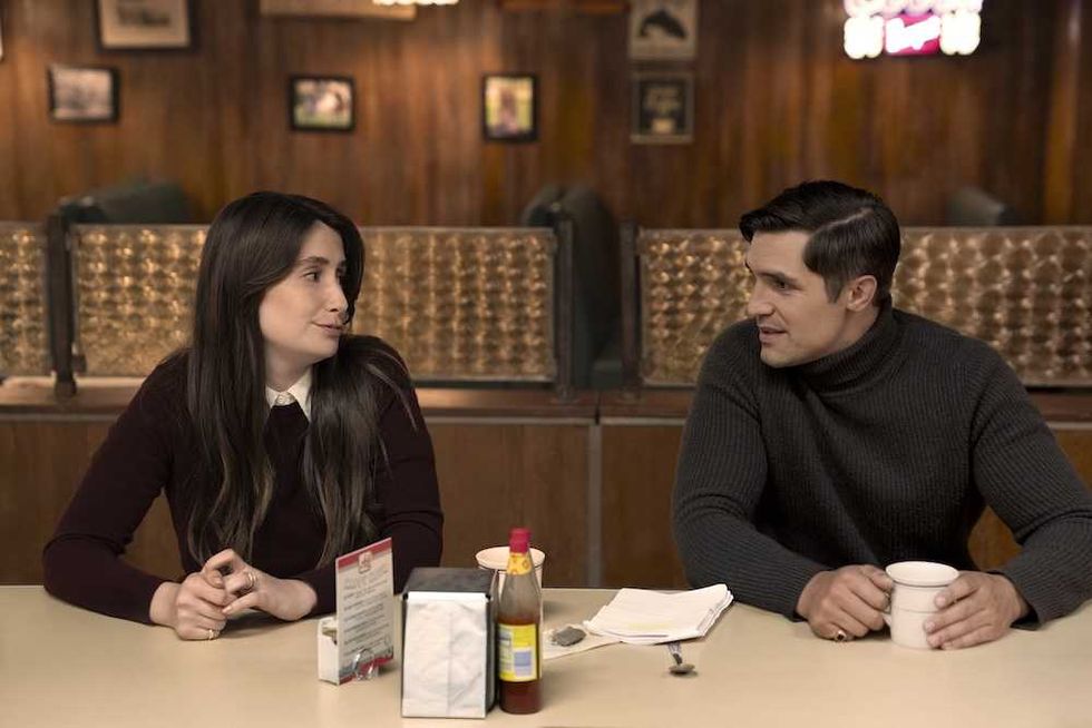 Two people converse at a diner counter with coffee cups and condiments in front of them.
