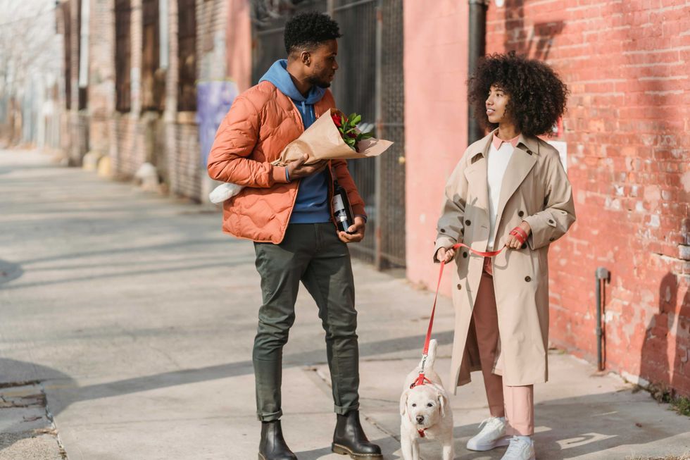 Two people converse on a sidewalk; one holds flowers, the other a dog on a leash.