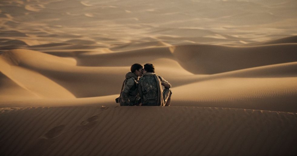 Two people crouch together on desert dunes, under warm light.
