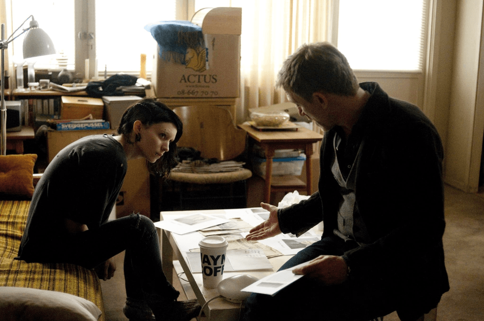 Two people discussing papers in a cluttered room with boxes and documents around them.