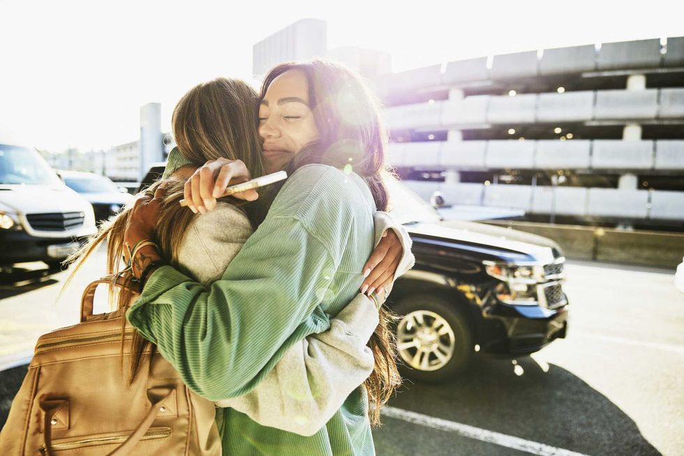 Two people embrace warmly in a sunny parking lot, one holding a phone.