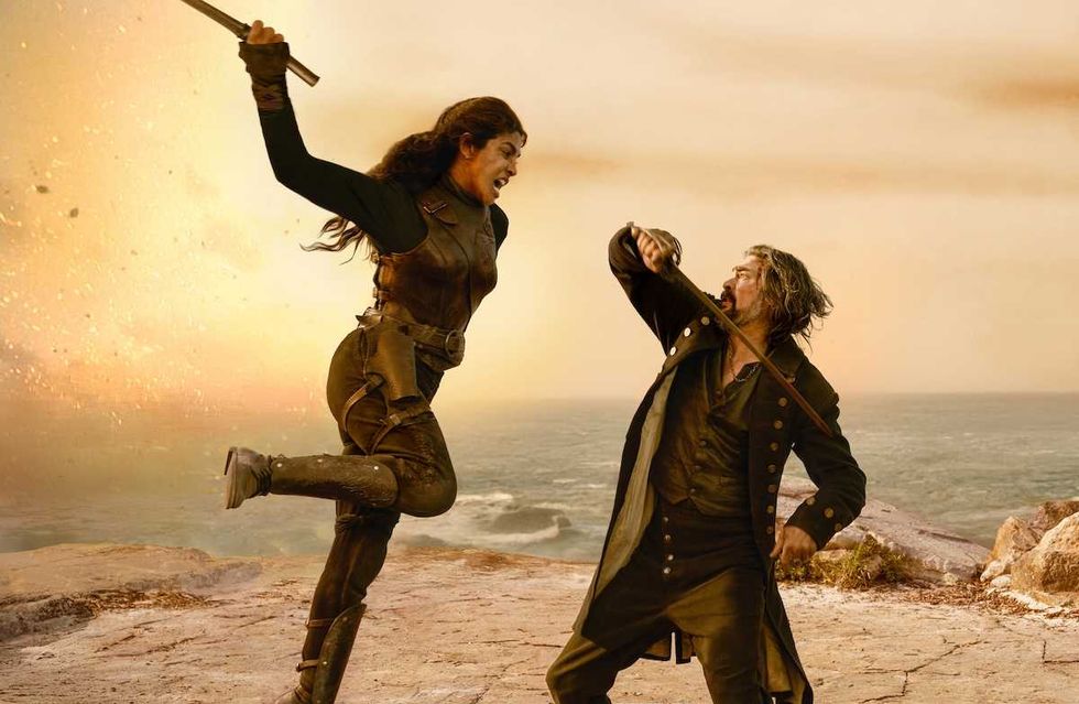 Two people engage in a dramatic sword fight by a seaside cliff at sunset.