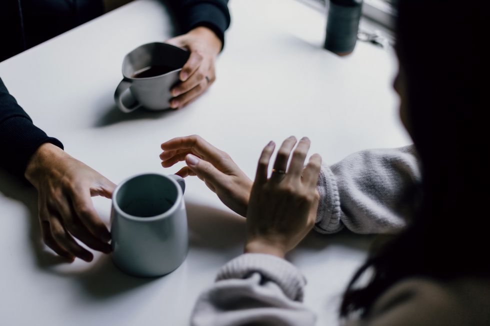 Two people having a conversation over coffee, hands resting on a white table.