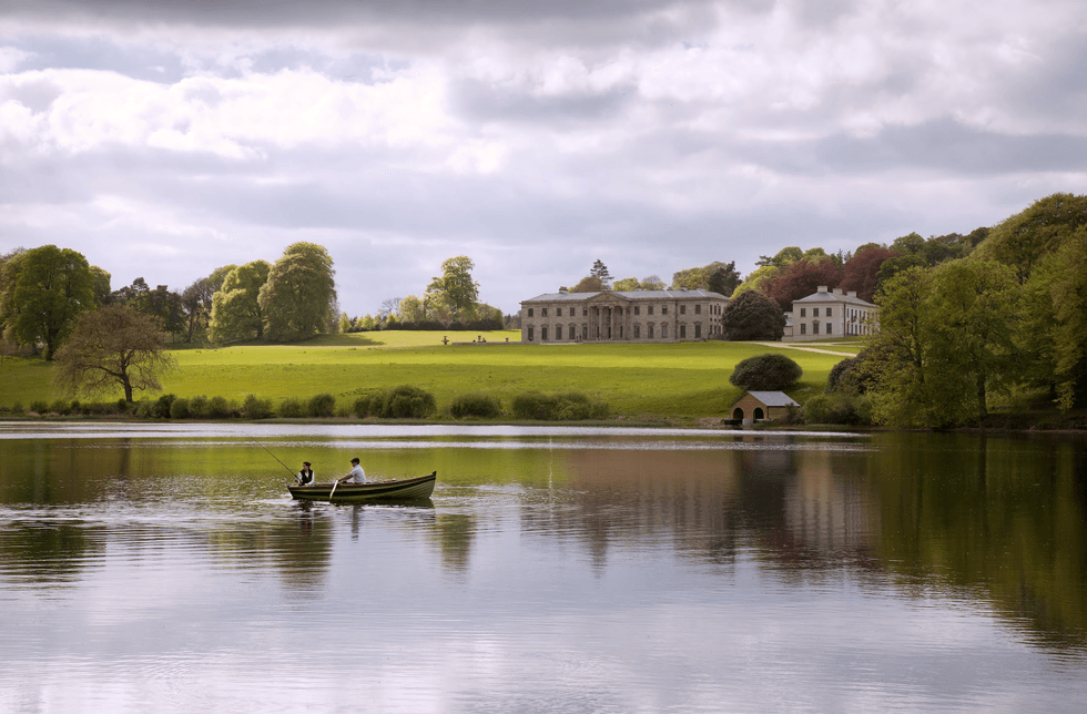 Two people in a boat fishing on a lake with a historic estate in the background.