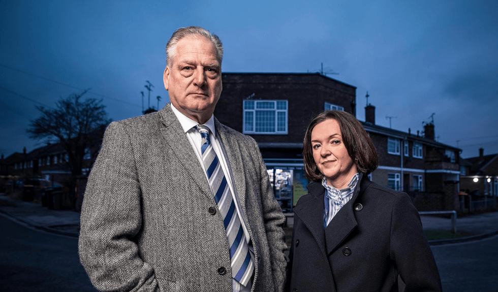 Two people in coats stand on a street at dusk with buildings in the background.