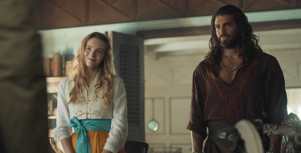 Two people in historical costumes stand smiling in a warmly lit room.