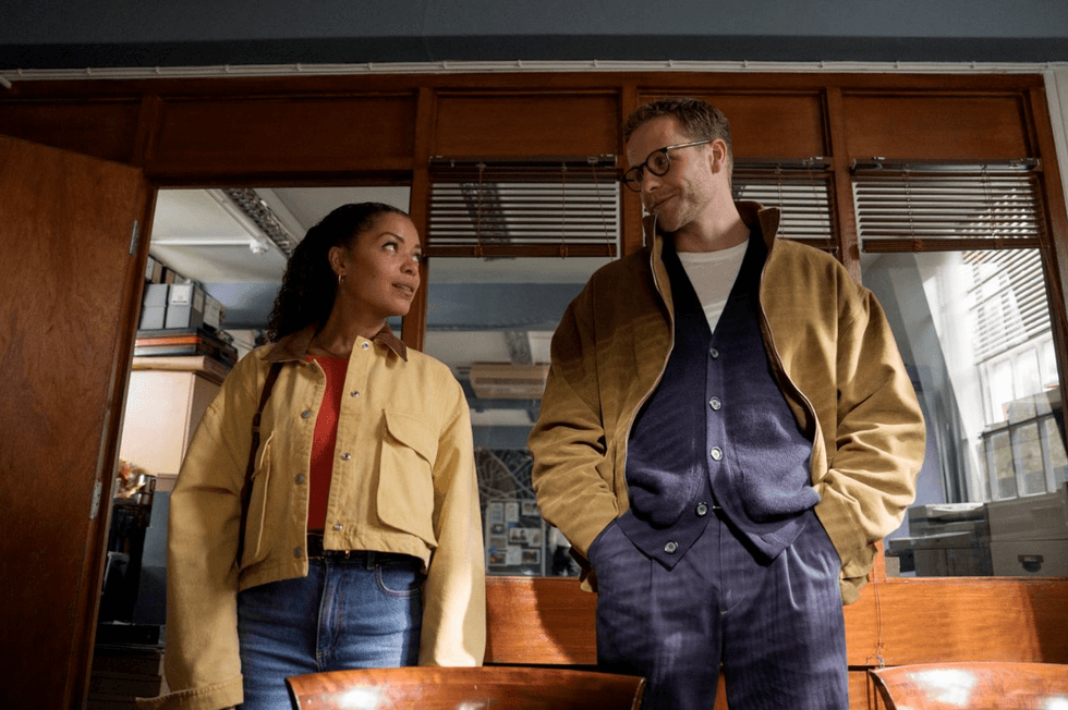 Two people in jackets stand in an office, looking at each other, with sunlight streaming in.
