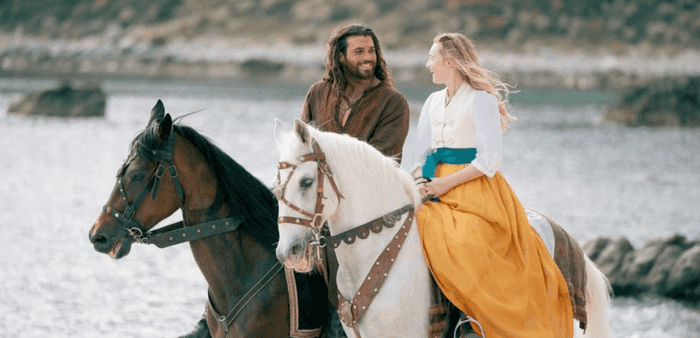 Two people in medieval-style clothing ride horses by the water, smiling at each other.
