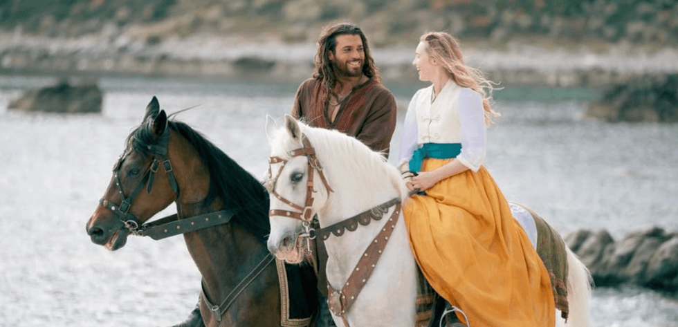 Two people in medieval-style clothing ride horses by the water, smiling at each other.