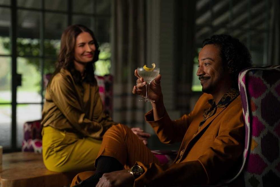 Two people in stylish outfits, one holding a cocktail, sitting indoors and smiling at each other.