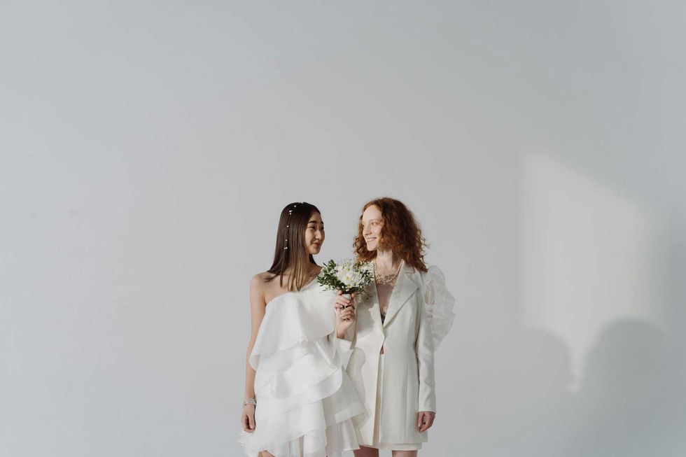 Two people in white attire smiling at each other, holding a bouquet against a plain background.