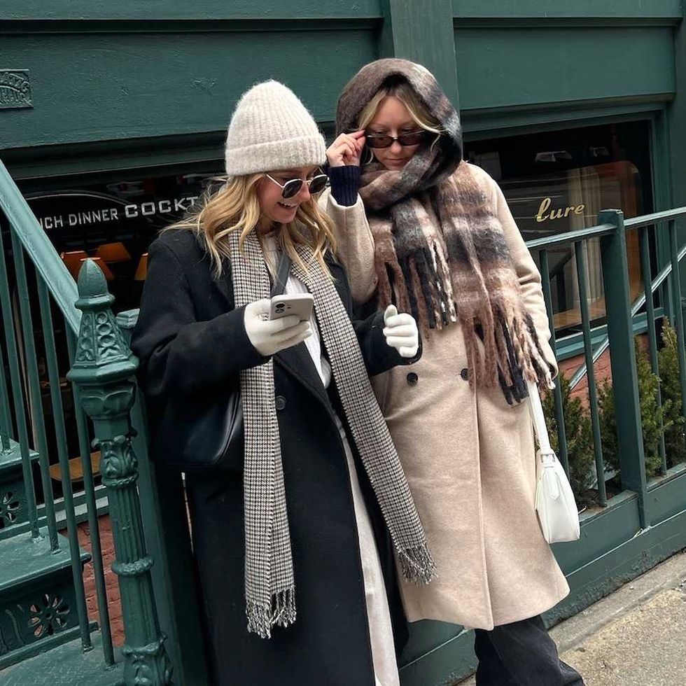 Two people in winter coats, scarves, and sunglasses, standing on a city sidewalk, smiling.