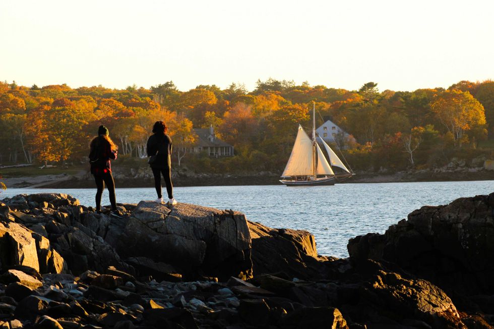 Two people on rocks watch a sailboat on a lake with autumn trees in the background.