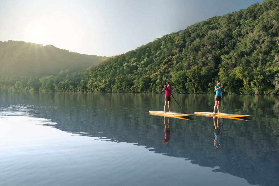 Two people paddleboarding on a calm lake surrounded by forested hills.