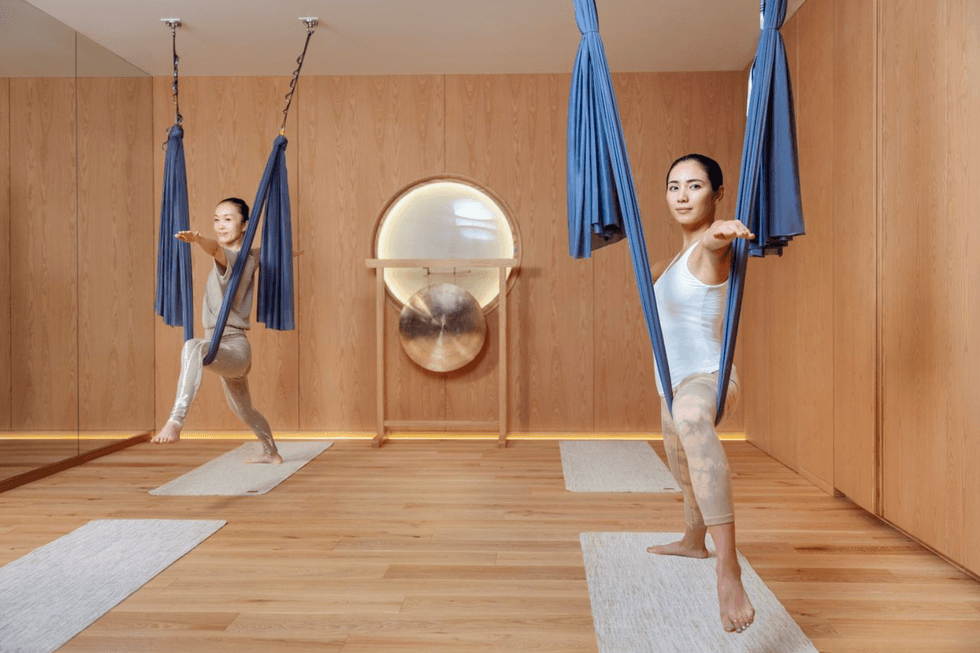 Two people practicing aerial yoga with blue hammocks, mirrored wall, and gong in the background.