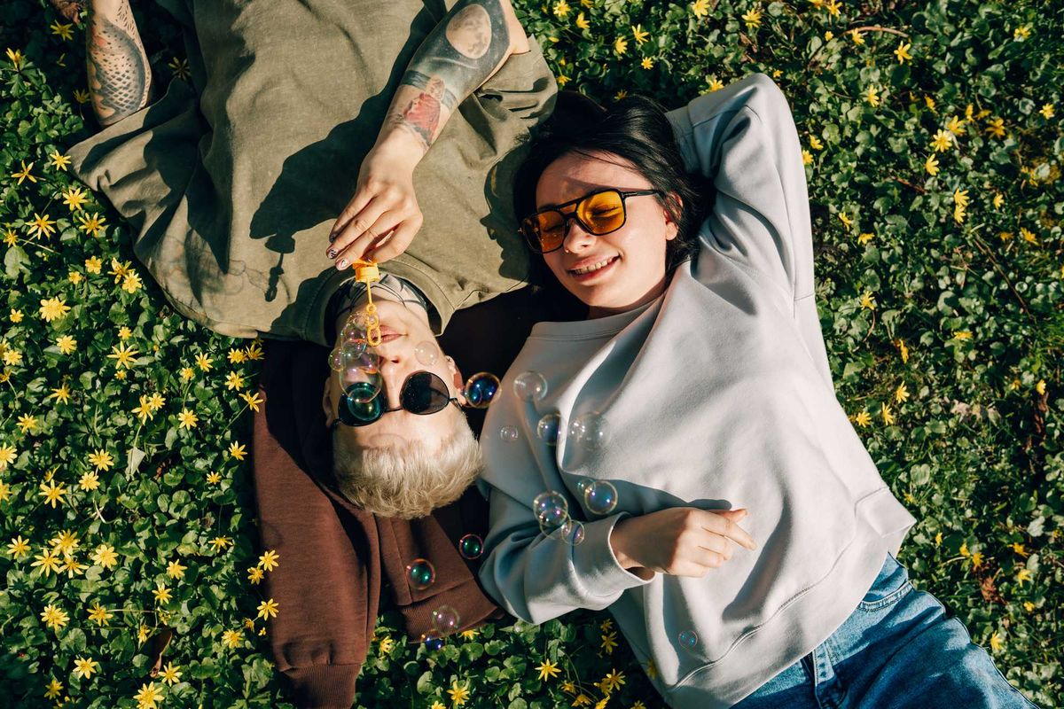 Two people relax in flowers, blowing bubbles, wearing sunglasses and casual clothing.