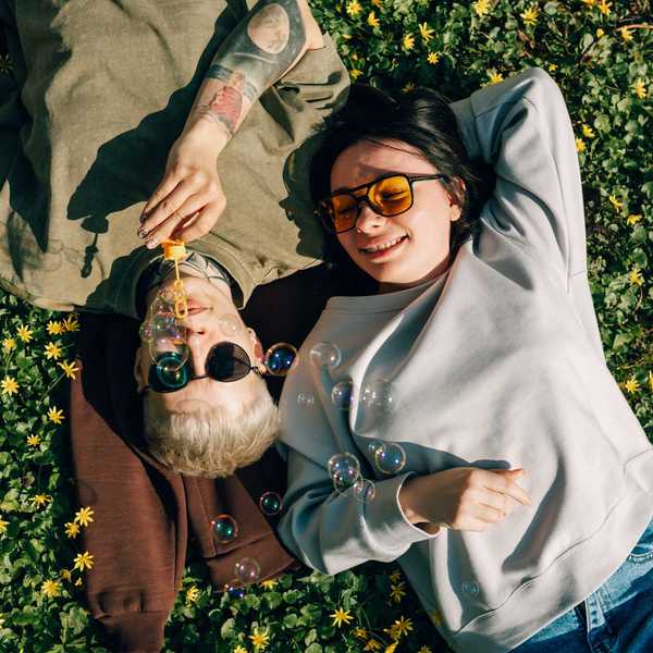 Two people relax in flowers, blowing bubbles, wearing sunglasses and casual clothing.
