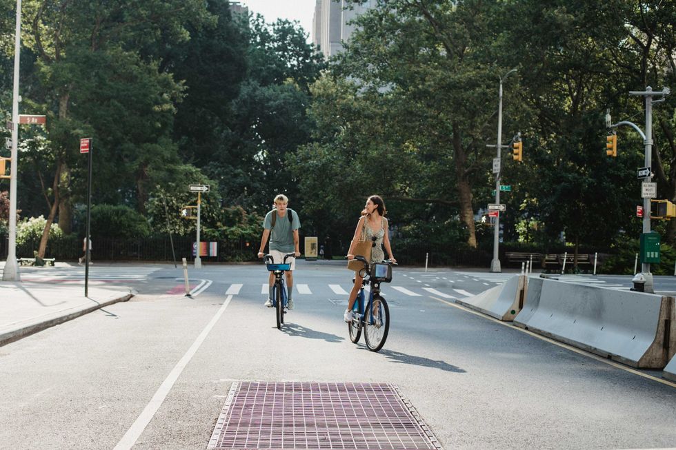 Two people ride bicycles on a sunny street with trees and traffic lights.