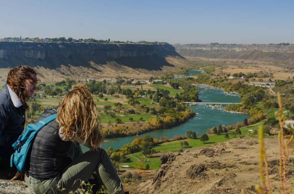 Two people sit on a cliff overlooking a river in a scenic canyon landscape.